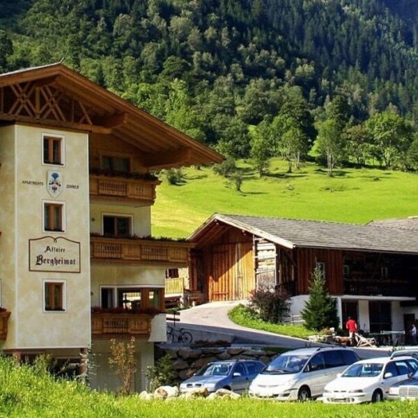 Vista esterna dell'agriturismo con balconi, un parcheggio e un parco giochi per bambini, sullo sfondo di montagne.