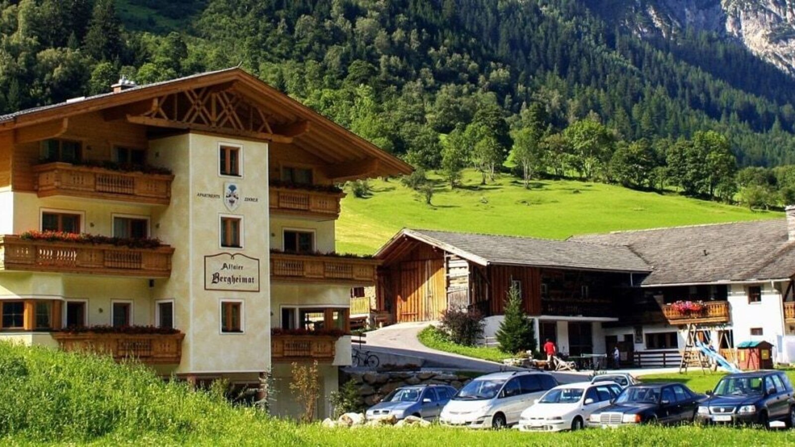 Vista esterna dell'agriturismo con balconi, un parcheggio e un parco giochi per bambini, sullo sfondo di montagne.