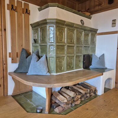 Una tradizionale stufa in maiolica verde nel agriturismo, con un'accogliente panca in legno, morbidi cuscini e legna da ardere ordinatamente accatastata.