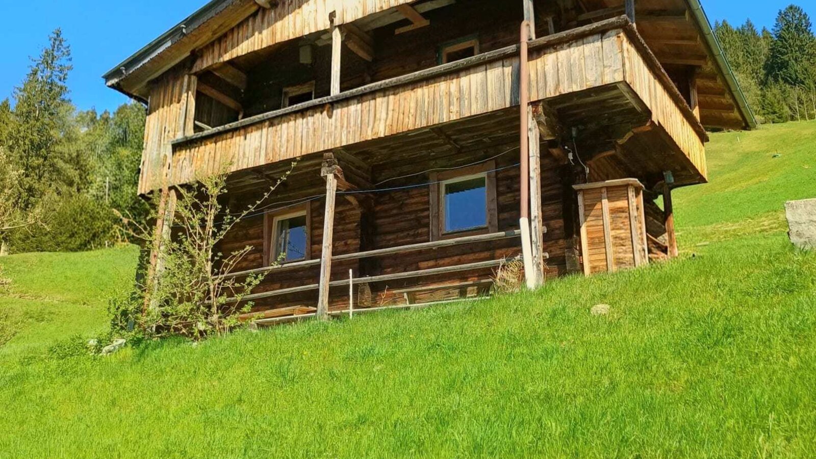 L'esterno dell'Alm, un edificio in legno a due piani con balcone su un pendio erboso.