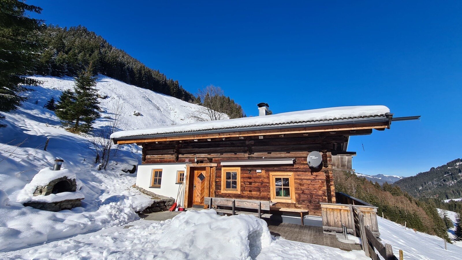L'Alm presenta una facciata in legno e un tetto innevato, circondata da montagne innevate sotto un cielo azzurro, con un forno esterno.