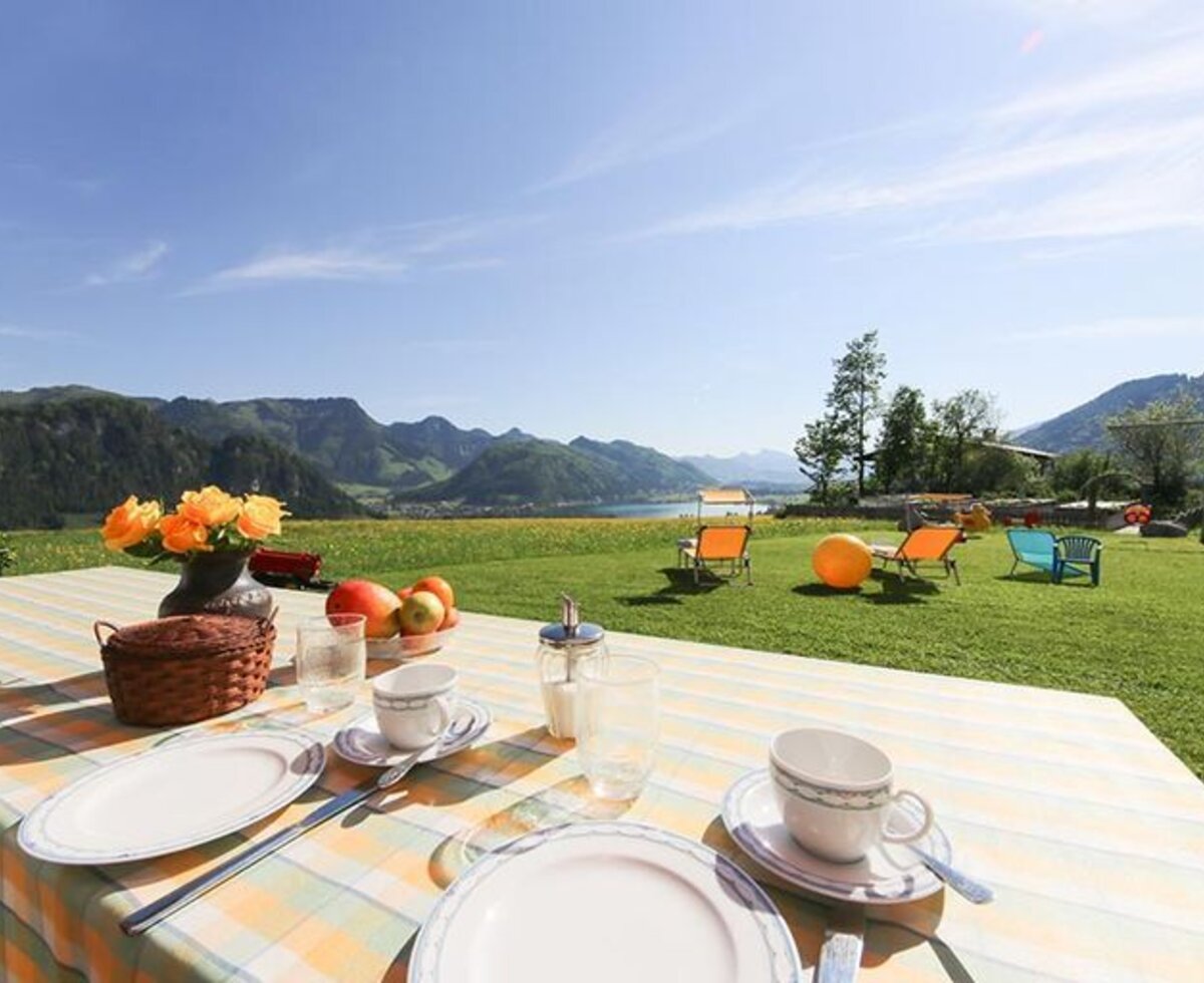 Tavolo per la colazione all'aperto nell'agriturismo con vista sul lago e sulle montagne, circondato da un prato con sedie a sdraio.