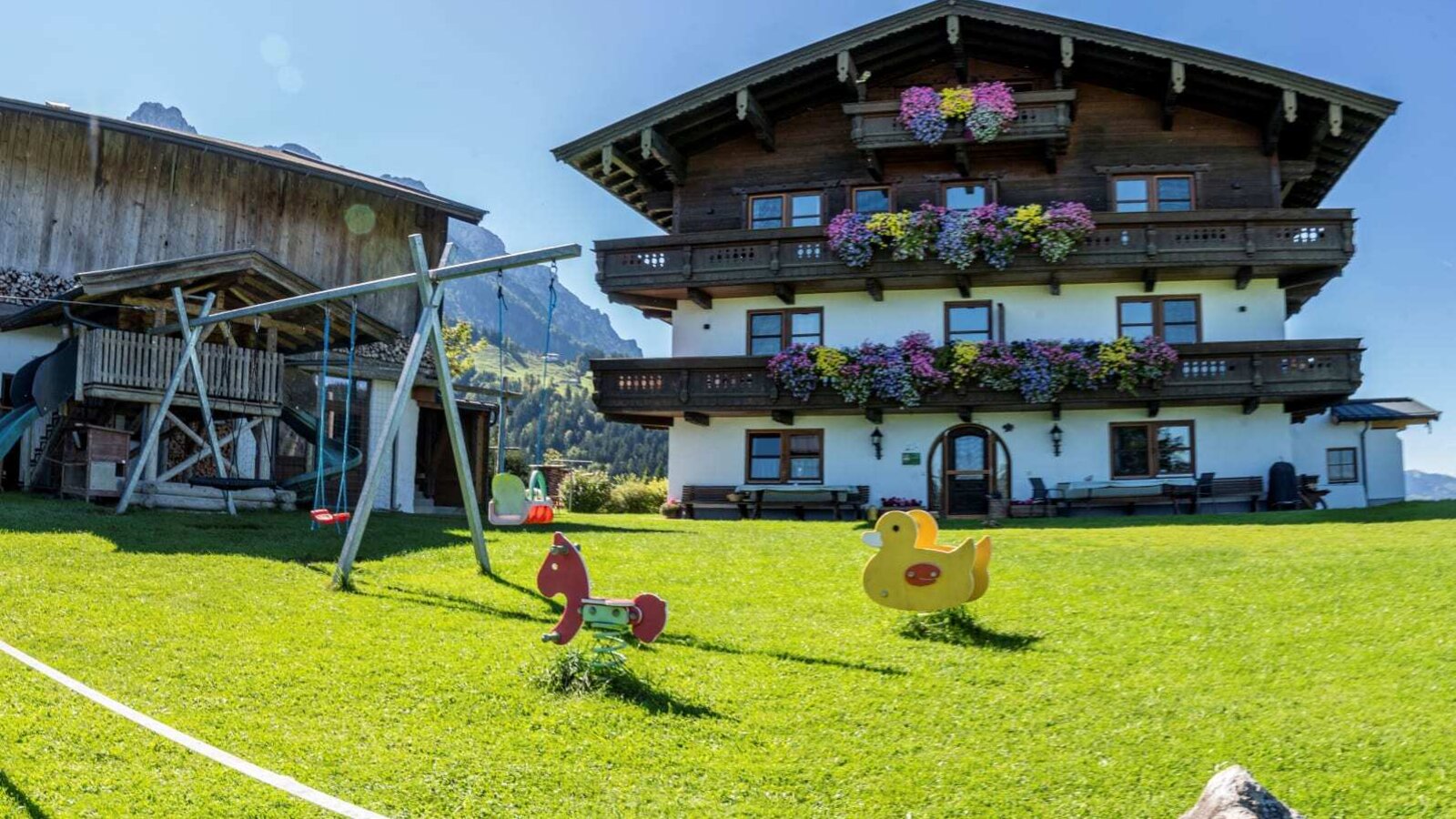 Vista esterna dell'agriturismo con balconi fioriti, un ampio prato con un'area giochi per bambini e una slackline.