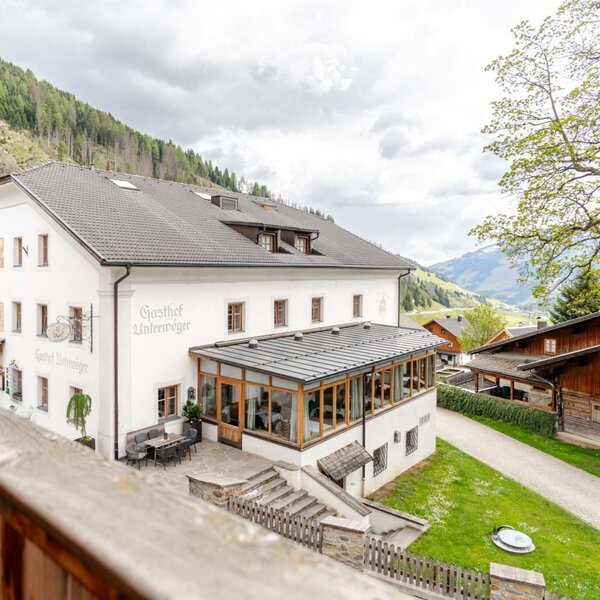 Vista esterna del Bauernhof, con un edificio principale bianco, una zona pranzo vetrata e una terrazza esterna, circondato dalle montagne.