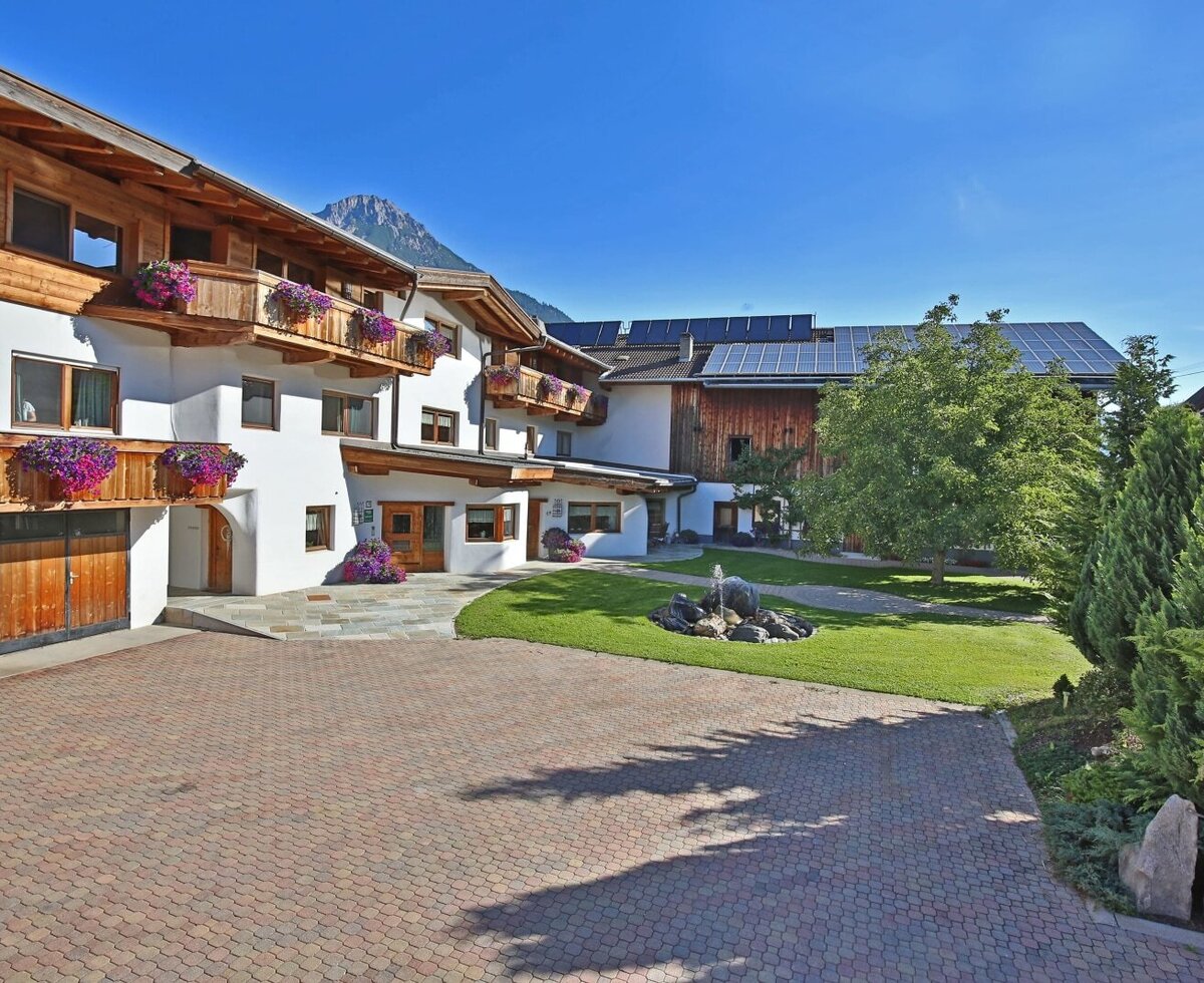 L'esterno dell'agriturismo, con un cortile pavimentato, un giardino con fontana, balconi in legno fioriti e vista sulle montagne.