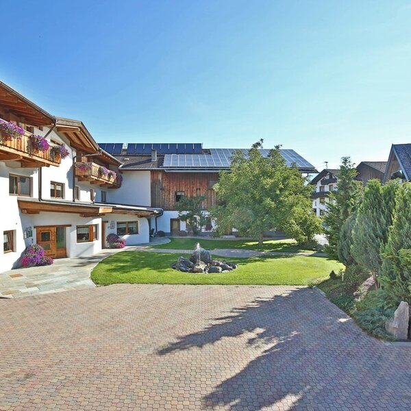 Vista esterna dell'agriturismo con balconi in legno fioriti, un cortile pavimentato e una fontana nel giardino.