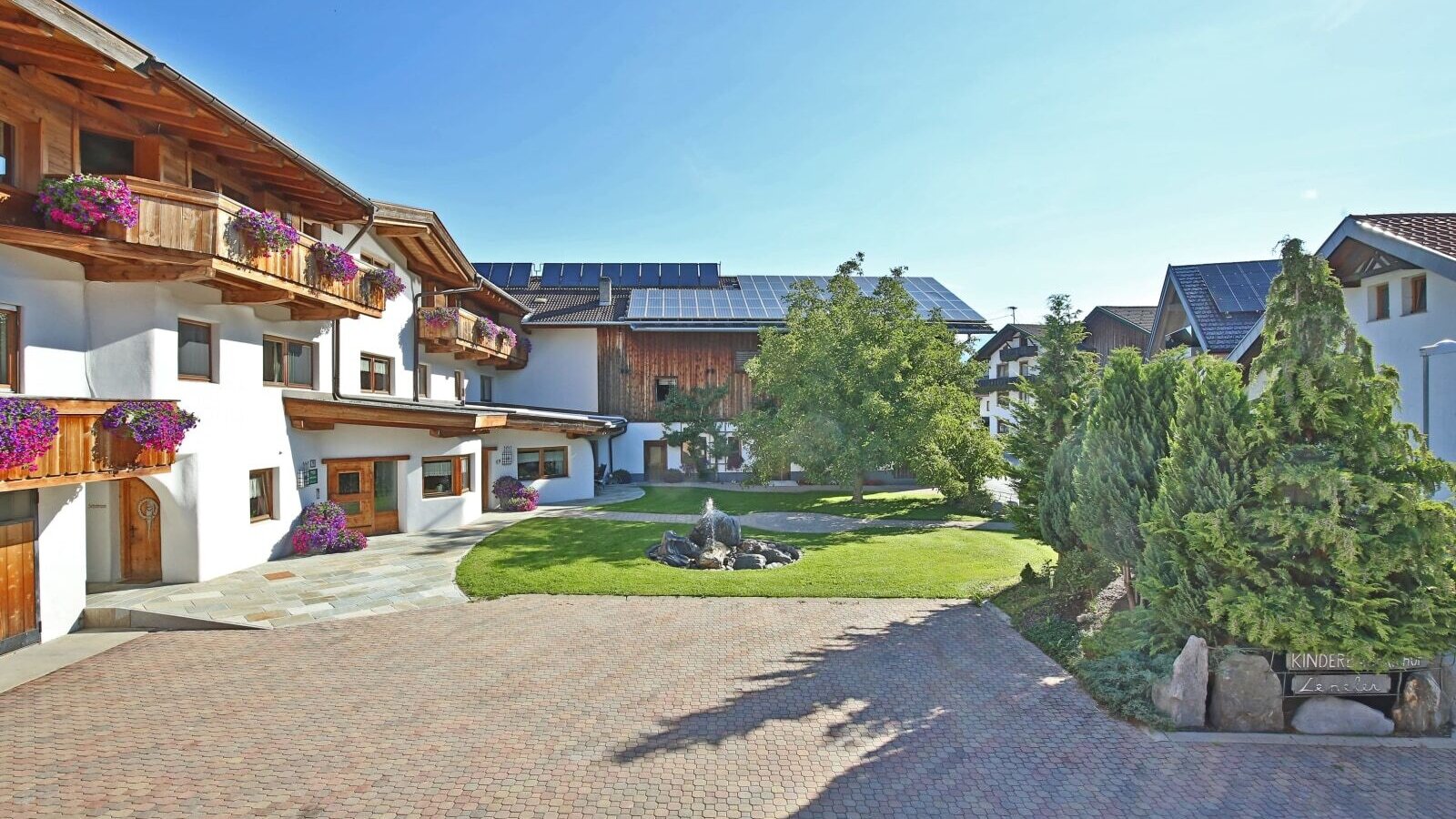 Vista esterna dell'agriturismo con balconi in legno fioriti, un cortile pavimentato e una fontana nel giardino.