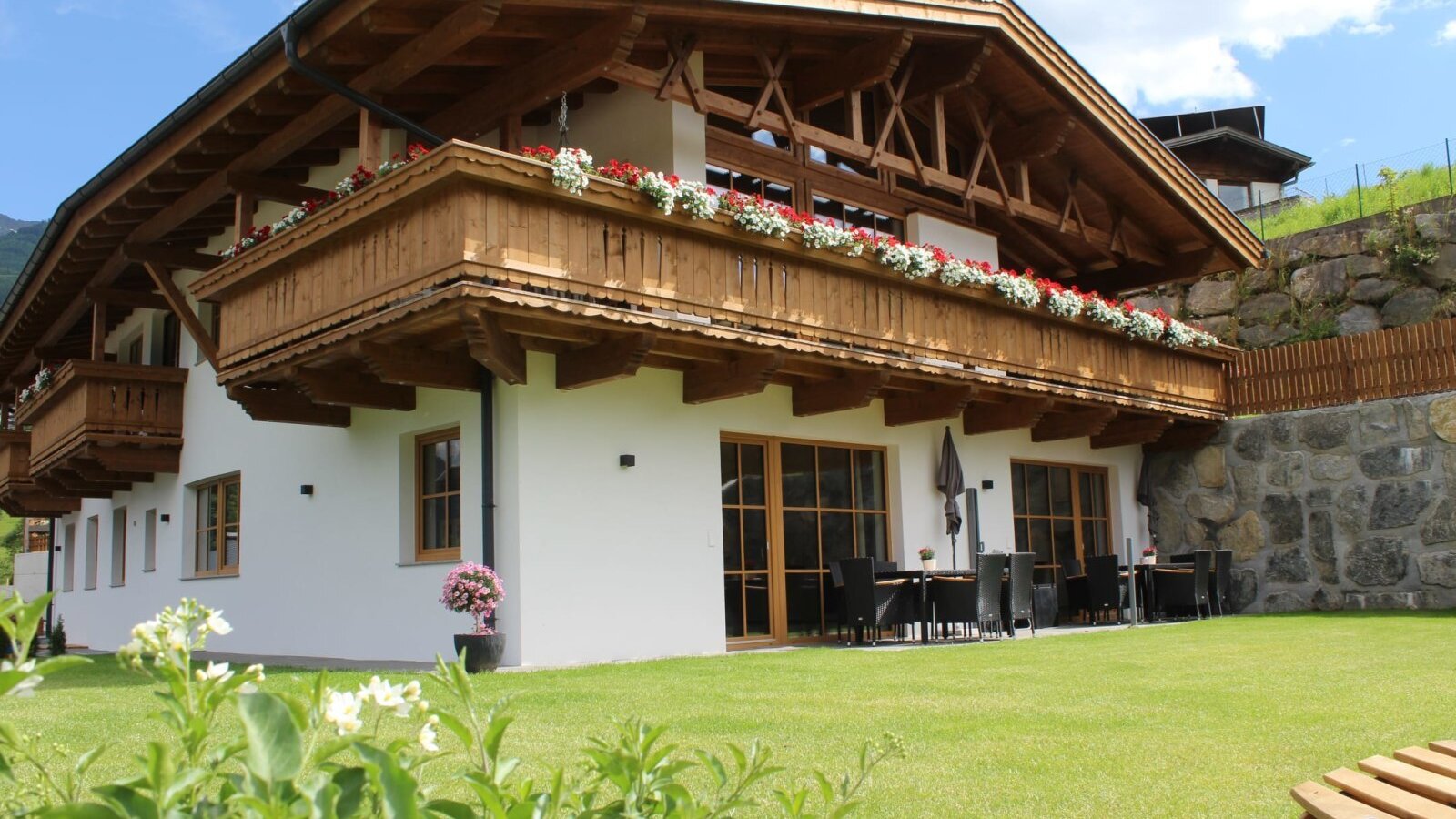 Vista esterna degli Ap­par­te­ments con balconi in legno fioriti, un prato verde e un'area salotto esterna.