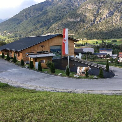 Vista esterna degli Ap­par­te­ments con facciata in legno, circondati dalle montagne, inclusa la strada di accesso, la bandiera austriaca e una statua di mucca.