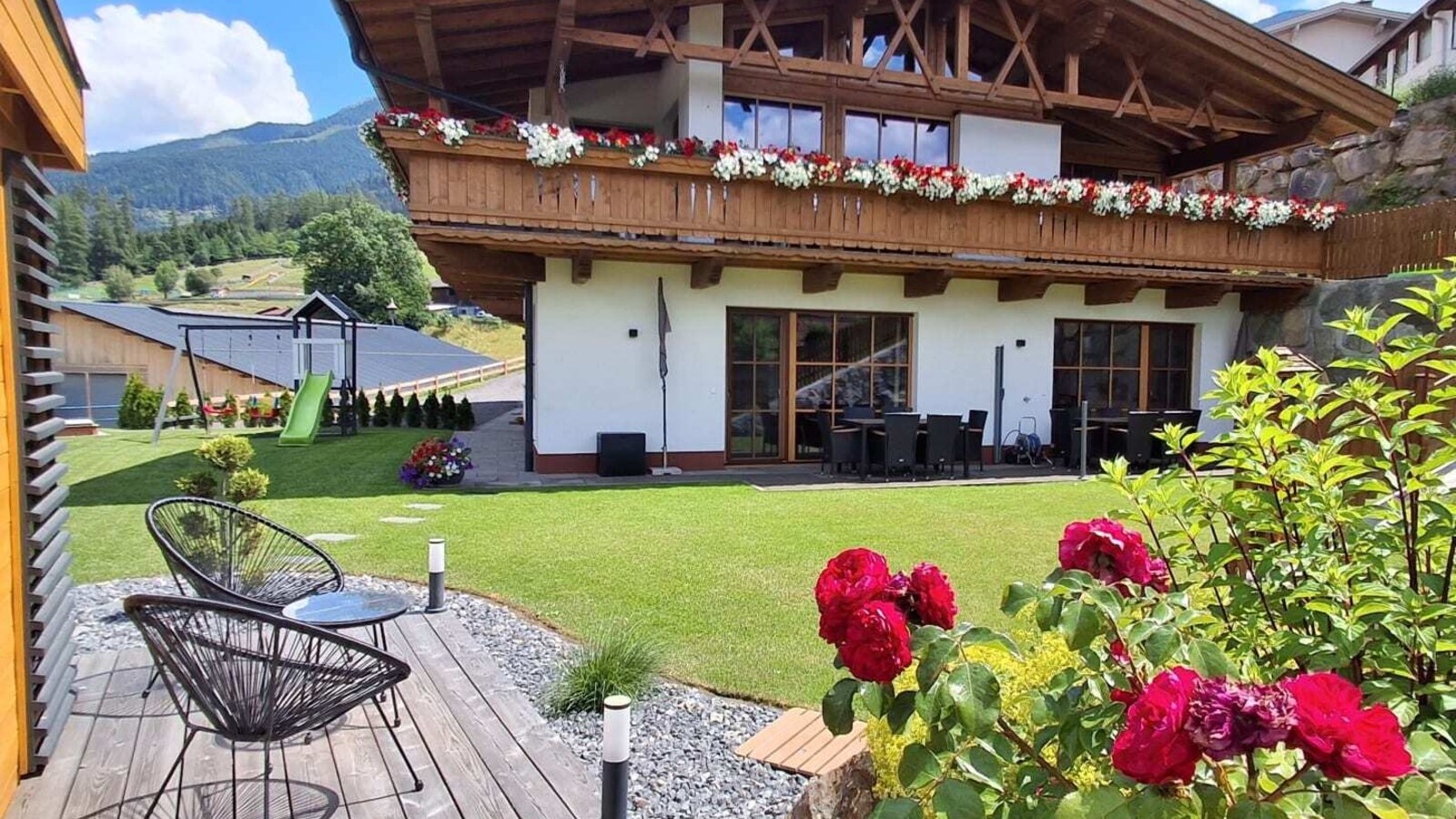 Esterno degli Ap­par­te­ments con balconi fioriti, giardino, area salotto esterna e parco giochi per bambini.