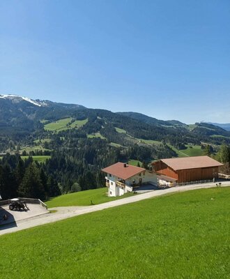 Vista esterna del Bauernhof con edificio agricolo adiacente, circondato da prati verdi e montagne.