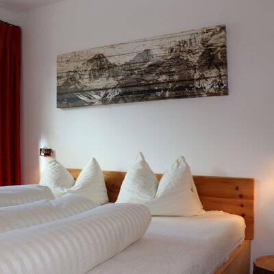 Un'accogliente camera da letto dell'appartamento con un letto in legno, soffici lenzuola bianche, un quadro alpino e calde luci da lettura.
